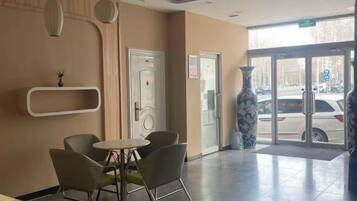 Un hall de hotel con mostrador de recepción, un área de descanso con sillas, un jarrón y una puerta que da al exterior.