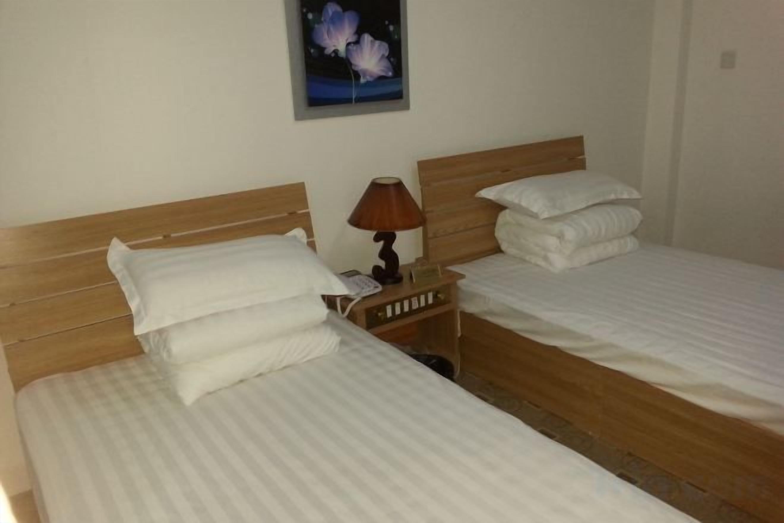 Quarto de hotel com duas camas, cada uma com roupa de cama branca e cabeceiras de madeira. Há uma mesinha de cabeceira com abajur entre as camas.