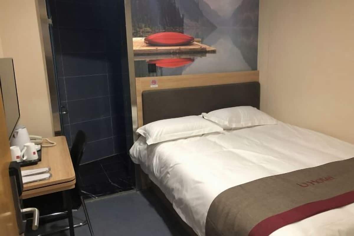 Une chambre d’hôtel avec un lit, un bureau et une chaise, un téléphone et une étagère fixée au mur sur laquelle se trouve un objet rouge.