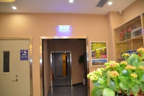 Un couloir intérieur avec une porte à gauche, un ascenseur au centre et une plante au premier plan.