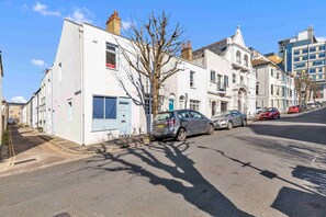 A street with parked cars and buildings, including a white building with a blue door and a tree in front. - Mill Cottage | By My Getaways (Brighton)