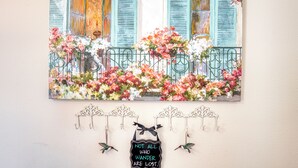 Ein Gemälde, das ein Gebäude mit blauen Fensterläden und einem Balkon voller Blumen zeigt. Darunter Wandbehänge mit einem Schild, auf dem steht: 'Not all who wander are lost.'