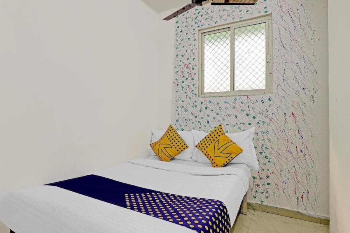 A small, single-bed room with a plain white wall and a window with a mesh screen. There are two pillows with a yellow and black pattern on the bed.