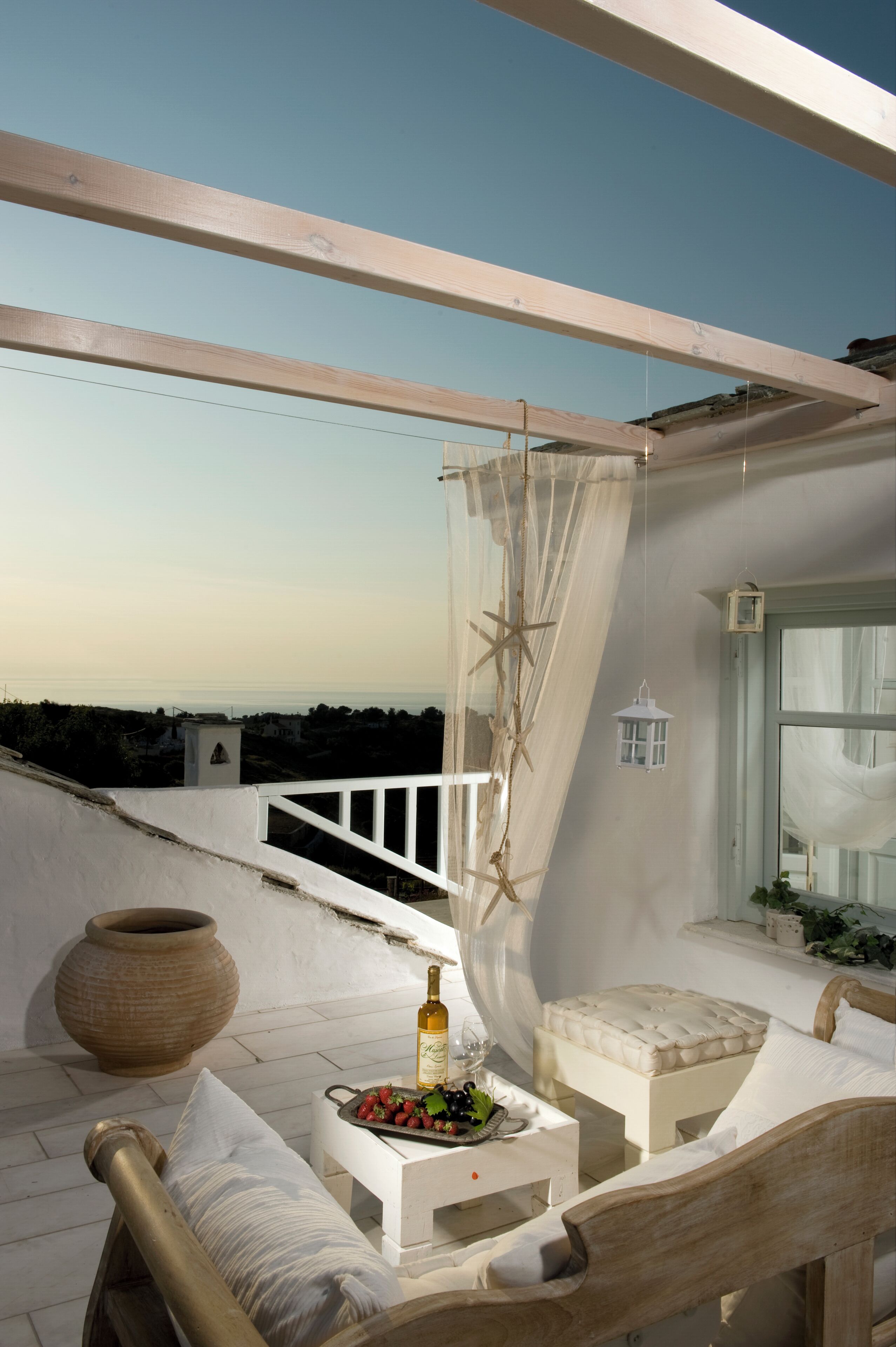 Un balcon avec une table blanche, dressée avec une bouteille, des verres et des fruits, un banc rembourré et une vue sur la mer.