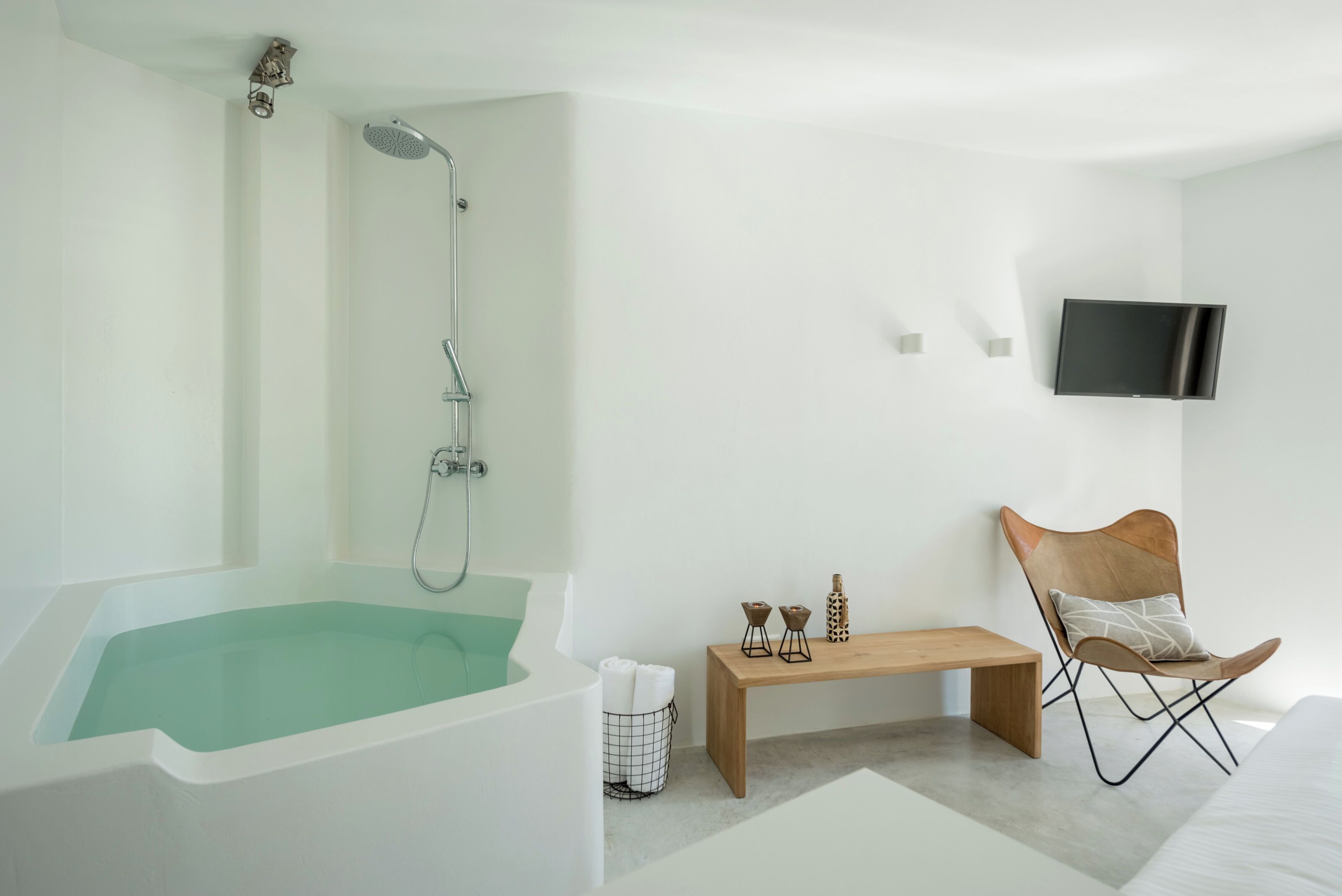 Ein modernes Badezimmer mit einer freistehenden Badewanne, einer Duschbrause, einer hölzernen Sitzbank und einem an der Wand montierten Fernseher.