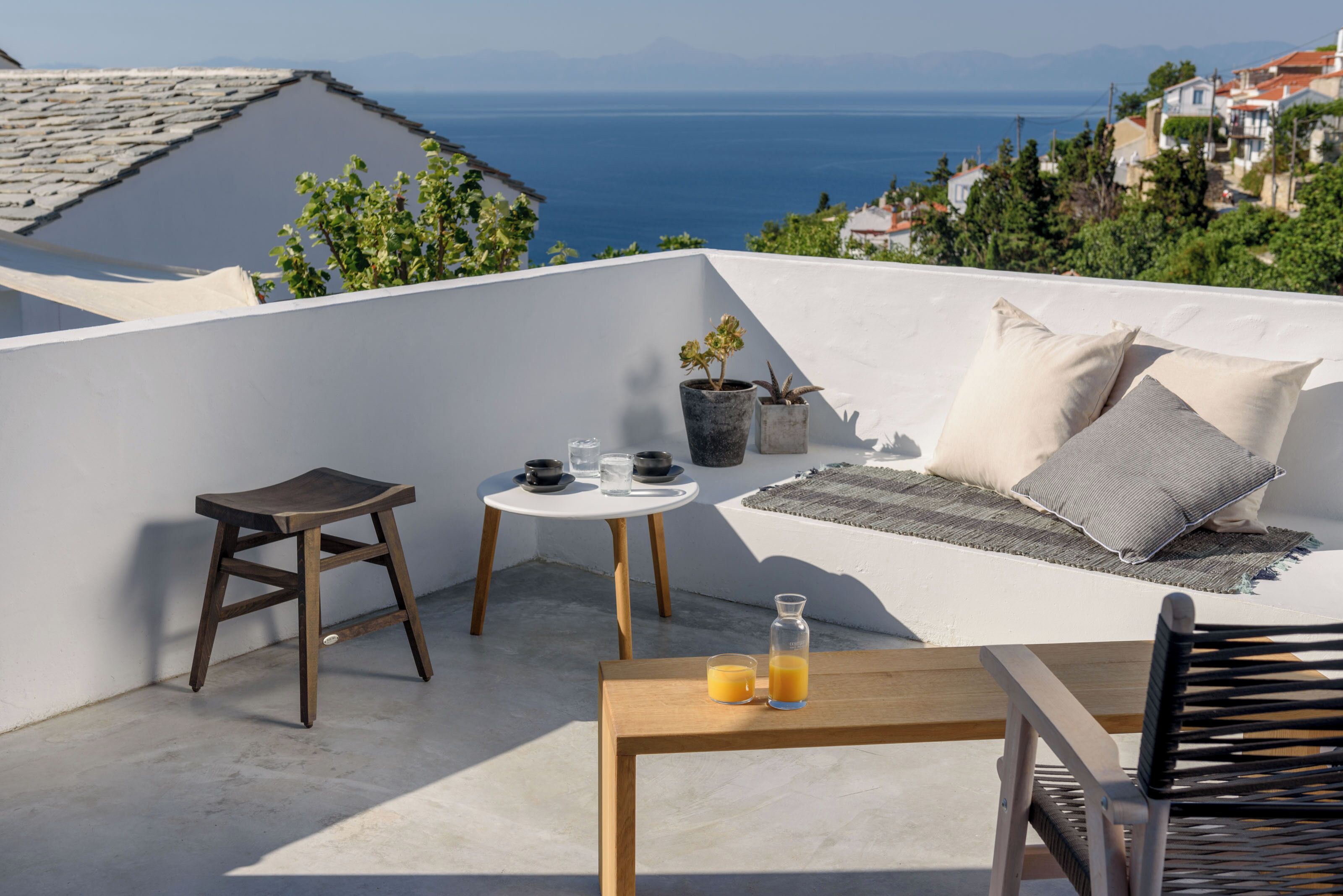Una terraza en la azotea con una mesa de madera, dos sillas, una mesita redonda con mantel blanco, algunas plantas en macetas y vistas al mar y edificios a la distancia.