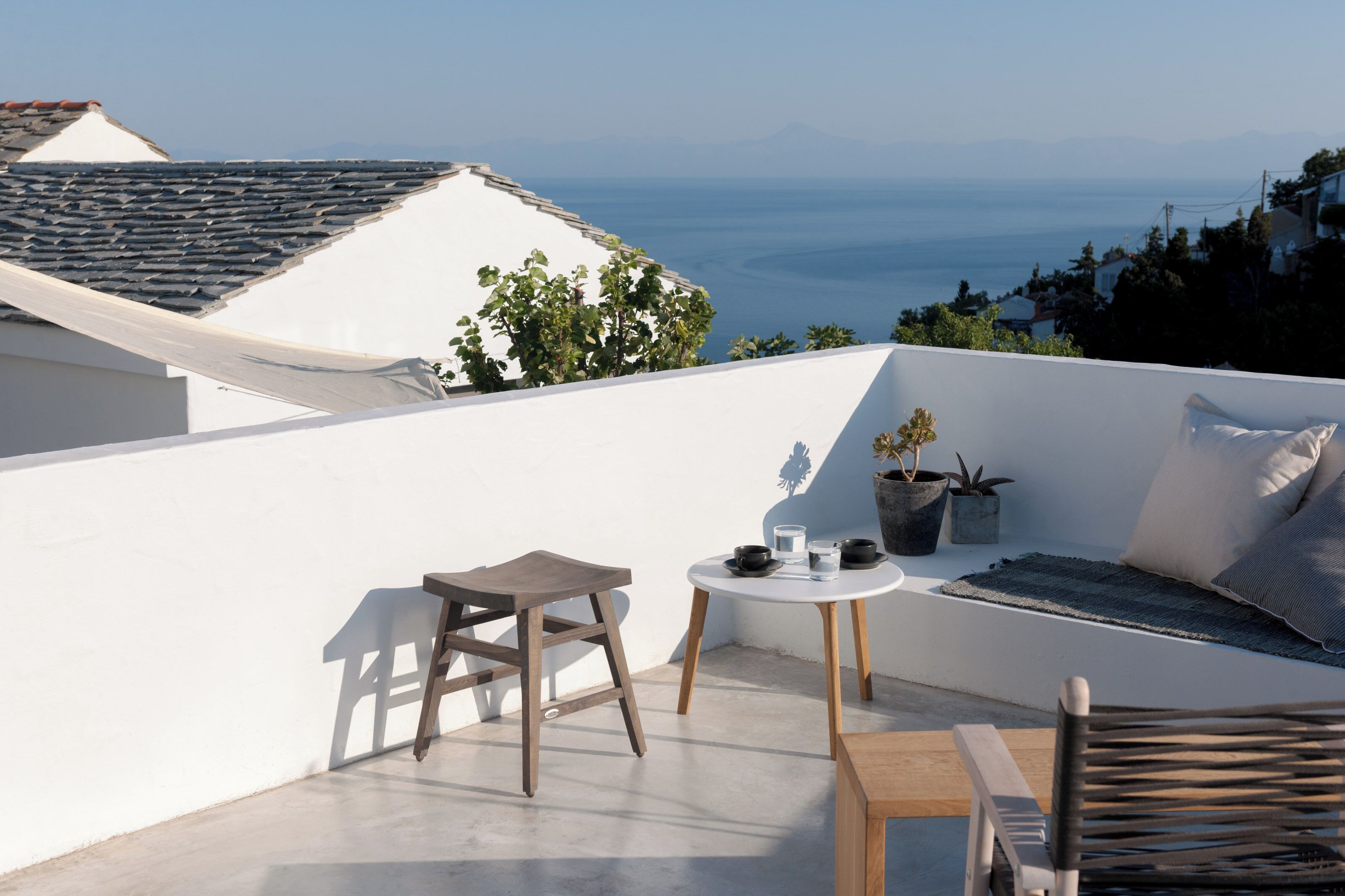 A rooftop terrace with a white wall, a small round table with a cup and saucer, a wooden stool, a cushioned seating area, and a view of the sea and distant mountains.