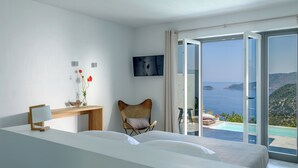 A modern bedroom with a large bed, a desk, a chair, and a TV. It features a balcony with a view of the sea.