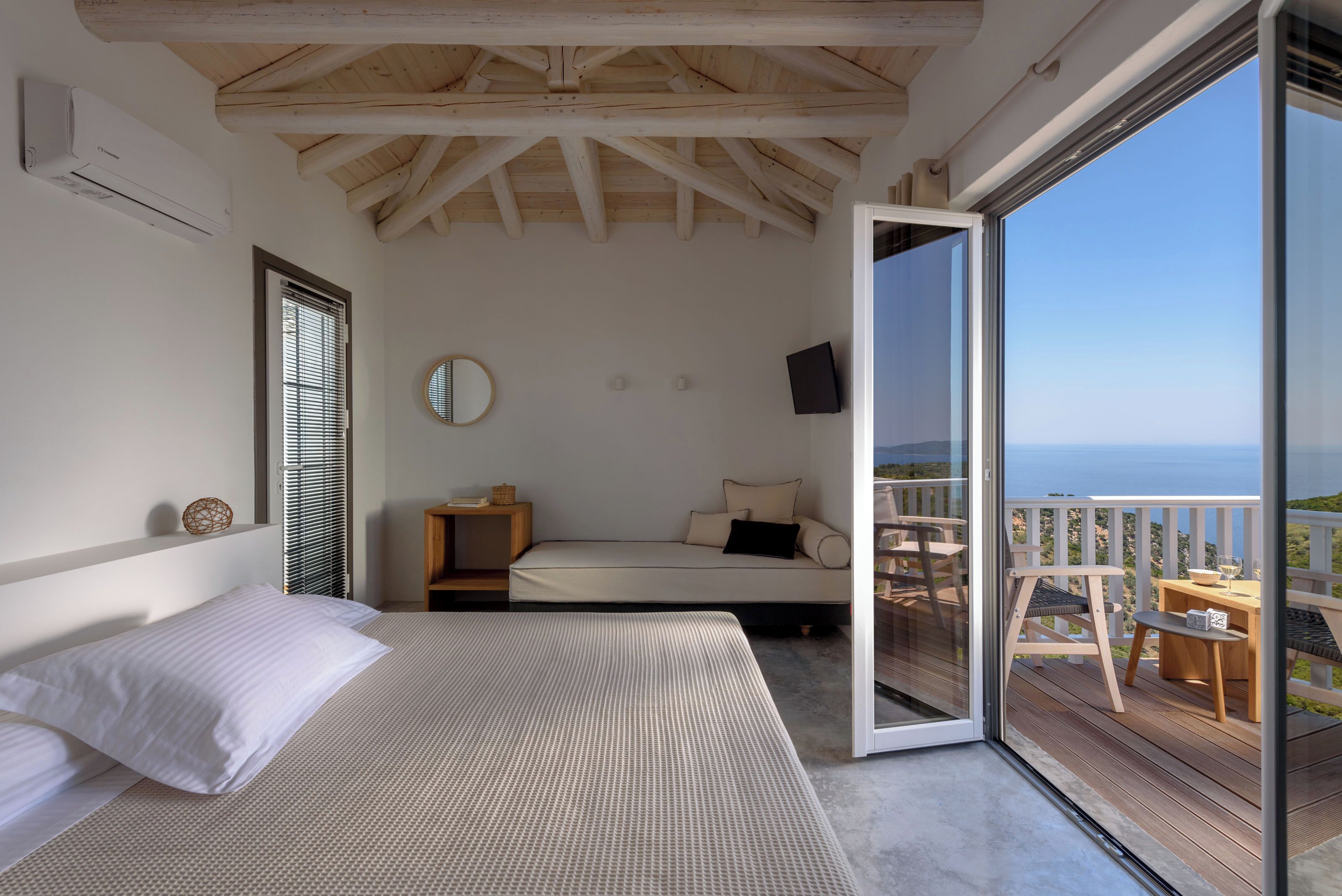 A bedroom with a bed, a bedside table, a mirror, and a sliding glass door leading to a balcony with a table and chairs.
