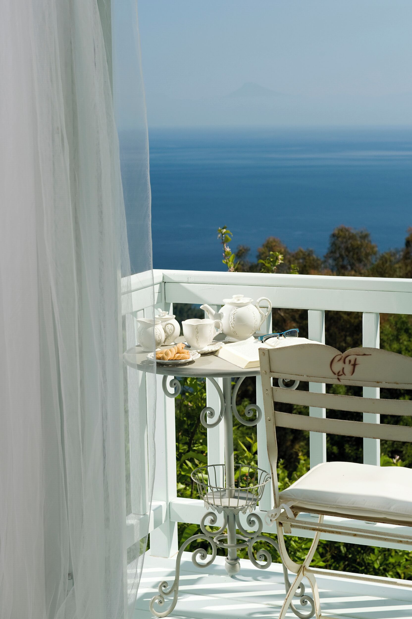 Un balcón blanco con vista al océano, una mesa con juego de té y una silla.