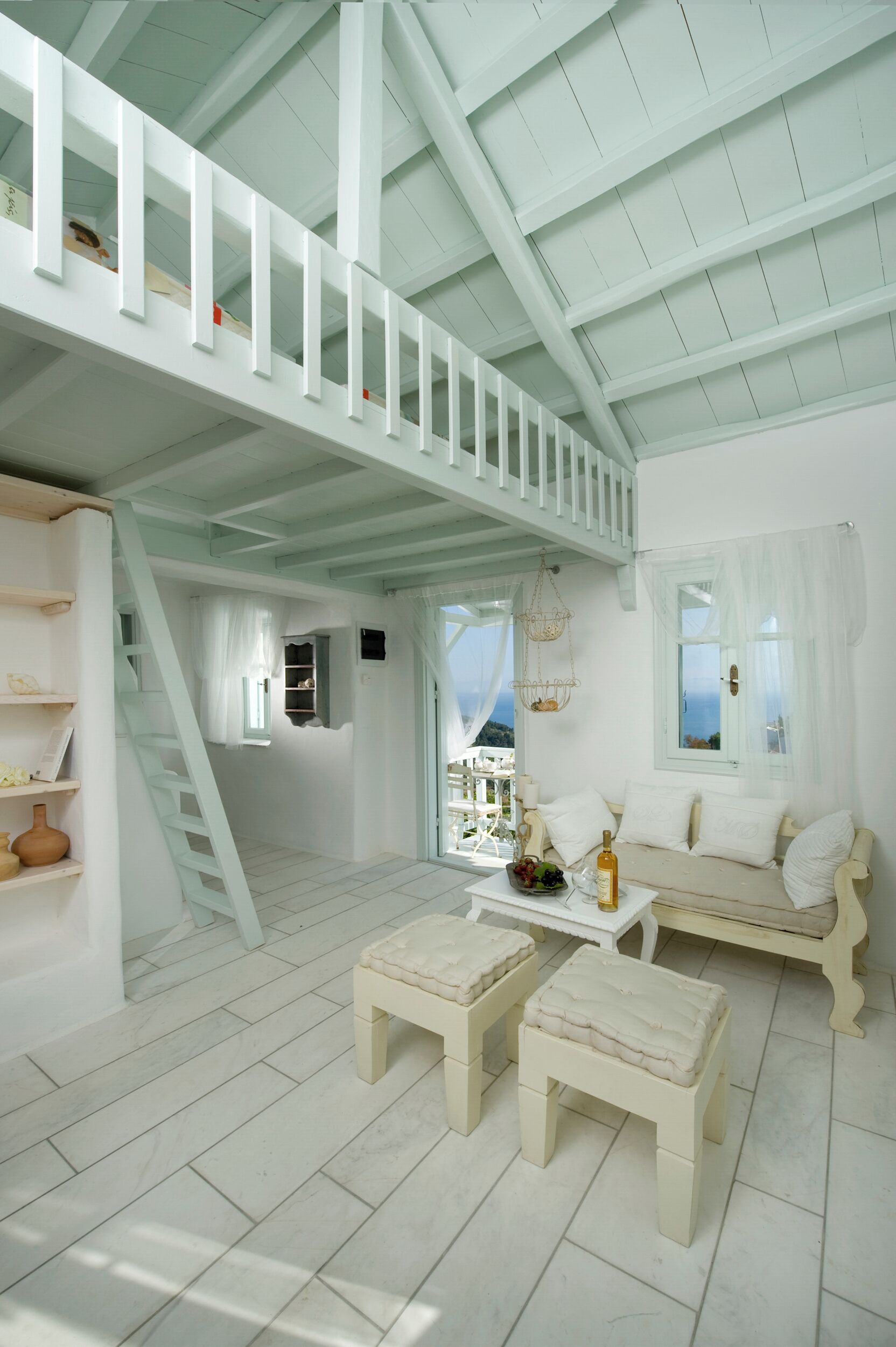 A spacious room with a high ceiling, white walls, and a staircase leading to an upper level. There is a seating area with a sofa and two ottomans, a small table with a bottle and fruit, and a view of the sea through a window.