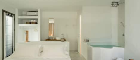 A modern bedroom with a large bed, a built-in wardrobe, a glass-enclosed bathtub, and a small wooden side table.