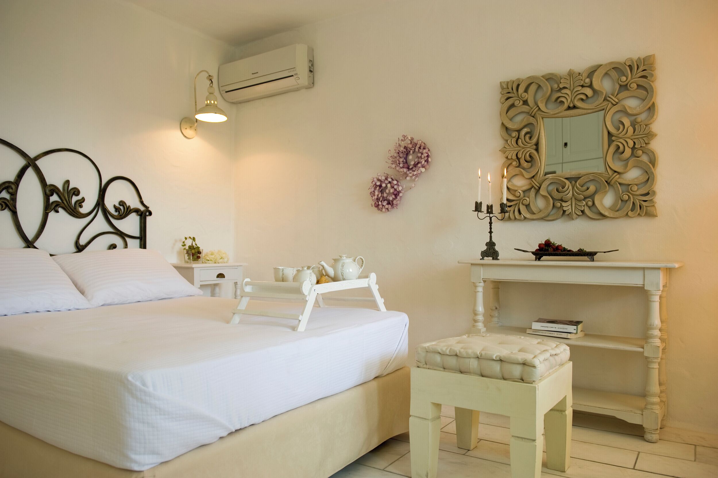 A bedroom with a bed, bedside table, lamp, mirror, and a small white bench.