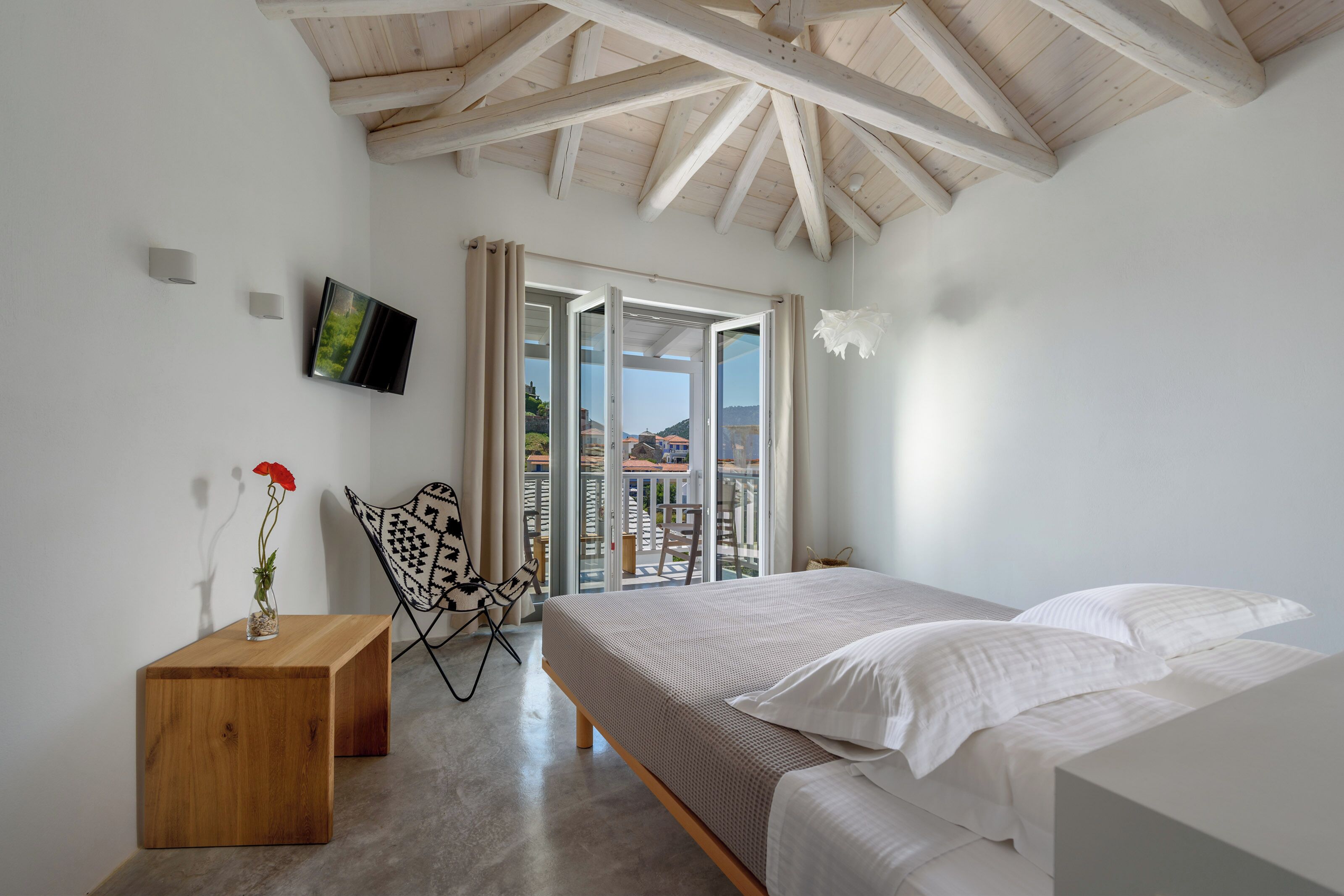 A bedroom with a bed, a wooden nightstand, a TV, a chair, and a balcony with a view.