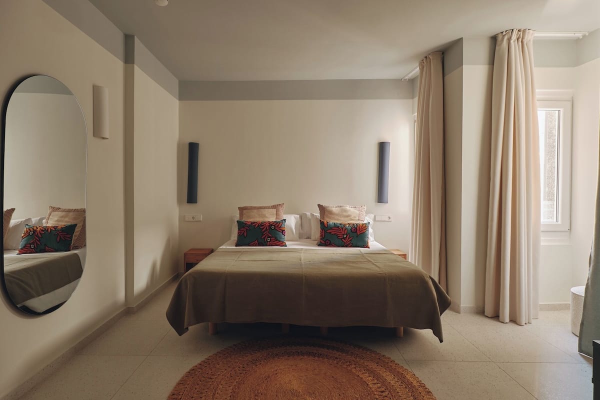 A bedroom with a large bed, a round mirror, a bedside table, a small nightstand, and a window with curtains.