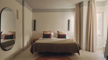 A bedroom with a large bed, a round mirror, a bedside table, a small nightstand, and a window with curtains.