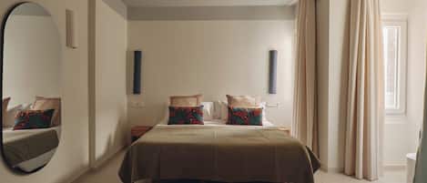 A bedroom with a large bed, a round mirror, a bedside table, a small nightstand, and a window with curtains.