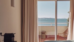 A hotel room with a bed, a desk with a coffee maker, a chair, and a view of the sea through a large window. - Atlante Sanxenxo Boutique (Sanxenxo)