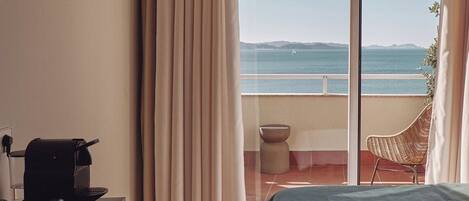 A hotel room with a bed, a desk with a coffee maker, a chair, and a view of the sea through a large window.