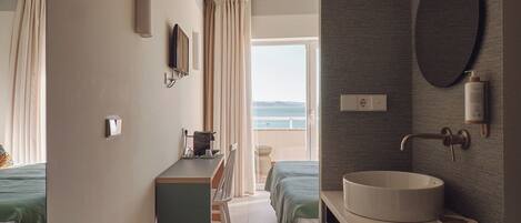 A hotel room with a bed, a desk, a bathroom with a sink, and a view of the sea.