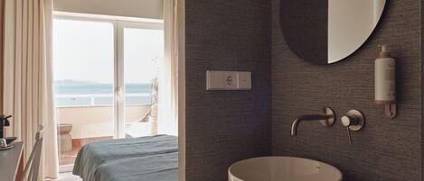 A hotel room with a bed, a round mirror, a sink, and a view of the sea through the balcony.