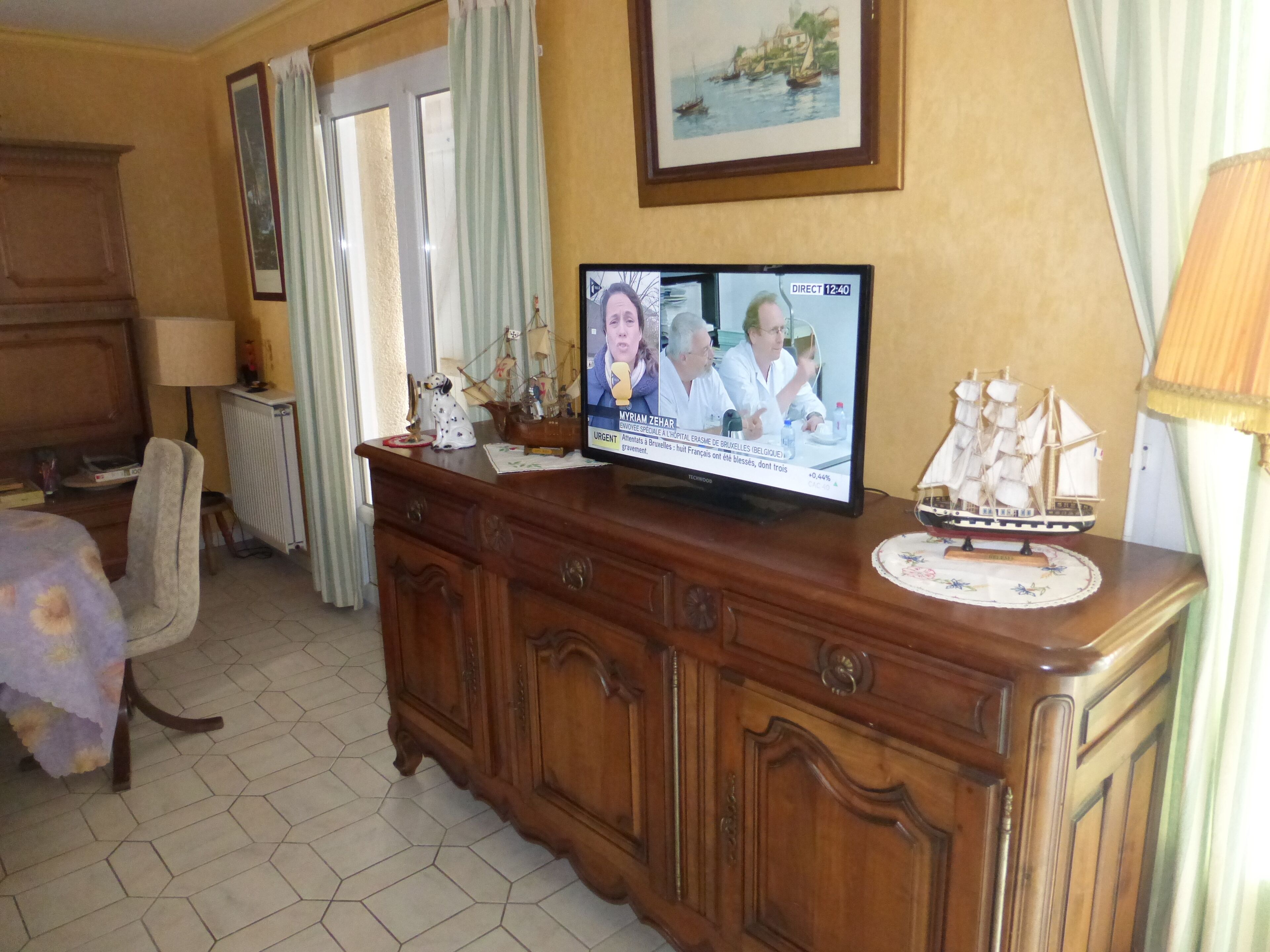 Une chambre d’hôtel avec une commode en bois, un téléviseur diffusant un programme d’information, une maquette de bateau et une fenêtre avec des rideaux.