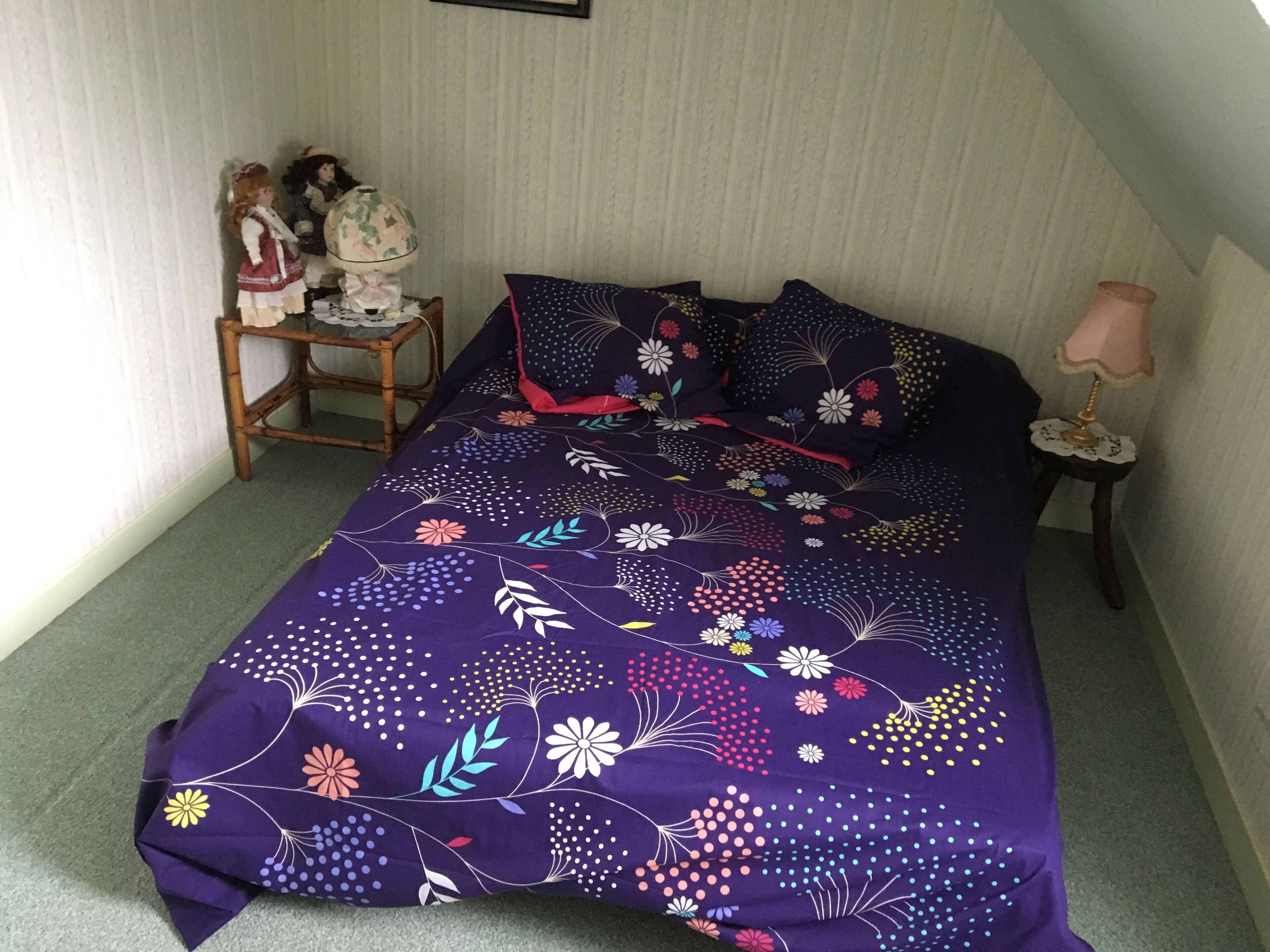 Un dormitorio con colcha de cama estampada en tonos morados, una pequeña mesa de noche de madera con lámpara, y una silla con una muñeca.