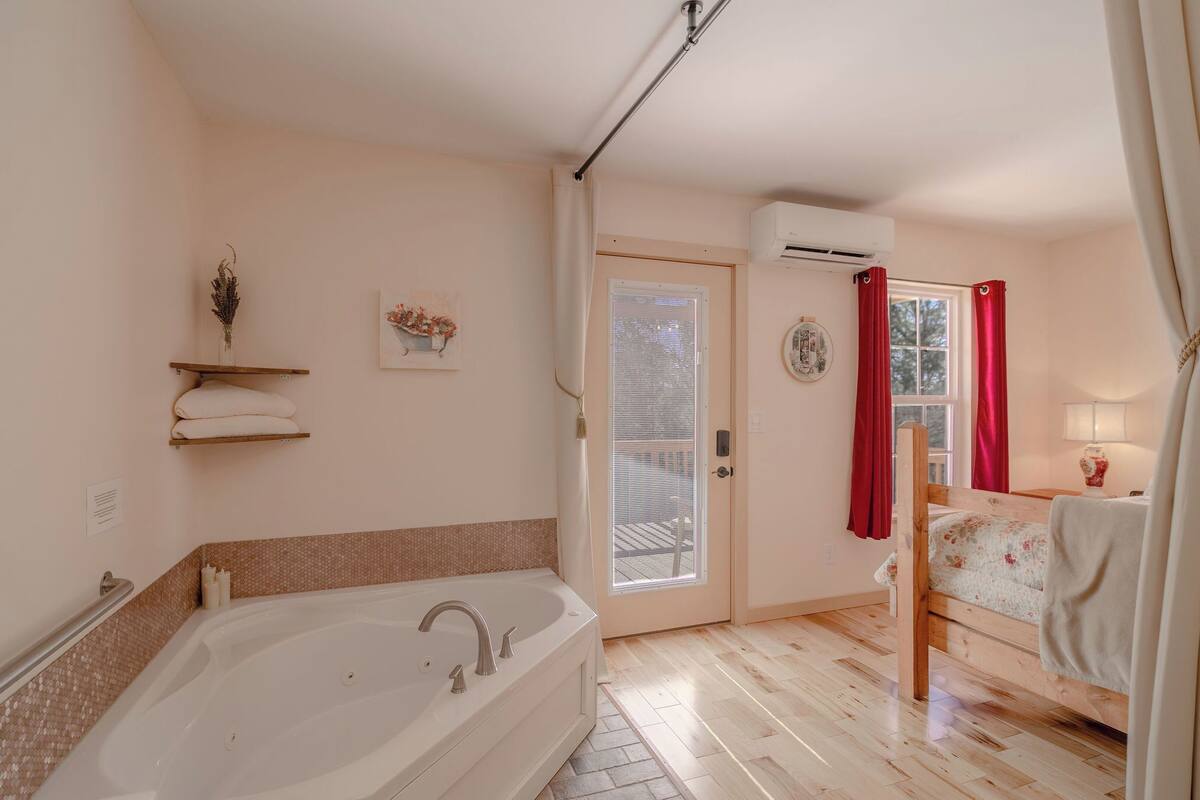 A bathroom with a bathtub, a shower, a toilet, and a sink. There is a window with red curtains, an air conditioning unit, and a towel rack.