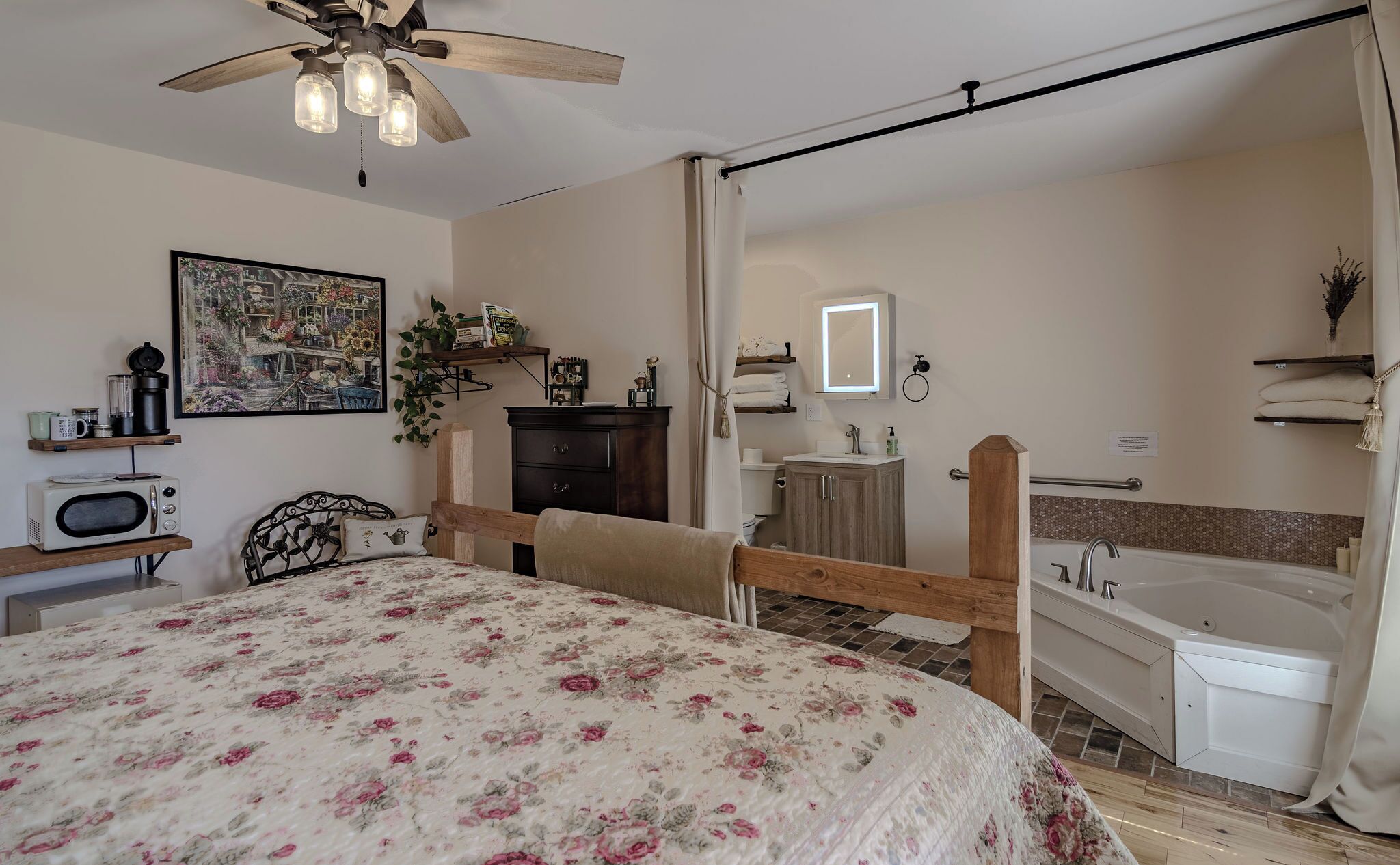 Un dormitorio con cama, ventilador de techo, mesita de noche, cómoda, baño con bañera, lavamanos y espejo.