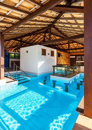 An indoor swimming pool with clear blue water, wooden beams, and a stone wall. - Bz16 Casa com Churrasqueira privativa (Buzios)