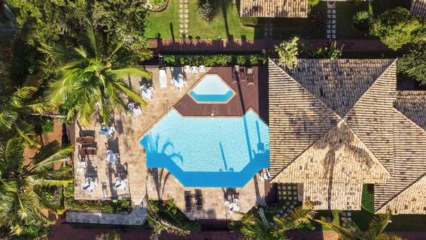 Vista aérea de un resort con una amplia piscina octagonal rodeada de sillas de descanso y sombrillas, palmeras y un edificio con techo de paja.