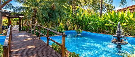 Terraza de madera que conduce a una piscina con una fuente, rodeada de exuberante vegetación y palmeras.