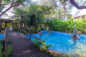 A wooden deck leading to a swimming pool surrounded by lush greenery and a decorative fountain. - Bz16 Casa com Churrasqueira privativa (Buzios)