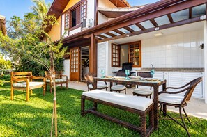 A patio with a dining table and chairs, a small garden, and a house with a balcony. - Bz16 Casa com Churrasqueira privativa (Buzios)