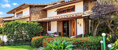 A two-story house with a stone facade, a tiled roof, and a well-maintained garden with red flowers and green shrubs.
