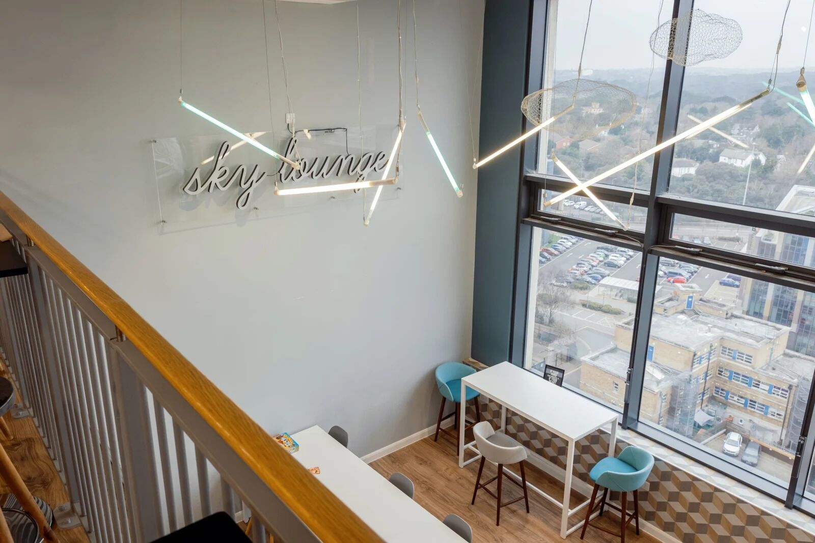 Ett modernt loungeområde med ett stort fönster som erbjuder utsikt över staden, ett vitt bord med barstolar och en neonreklamskylt med texten "Sky Lounge".