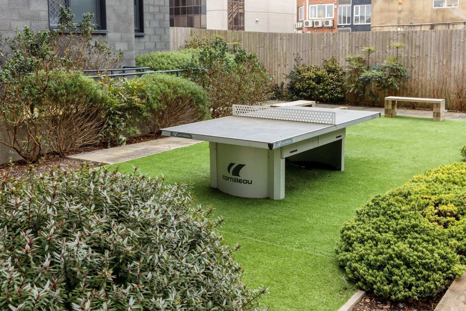 Mesa de ping pong en un jardín bien cuidado con césped artificial, rodeada de arbustos variados y una valla de madera.