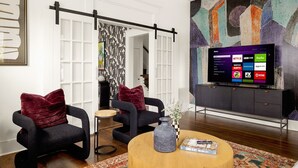 A modern living room with a flat-screen TV, two black armchairs with red cushions, a round coffee table with a vase and a book, and a colorful abstract painting on the wall. - La Flor: Family Dream Home ~ .5mi to Pearl ~ Pool (San Antonio)