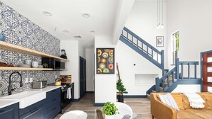 Eine moderne Küche mit dunklen Schränken, einer weißen Arbeitsplatte und einer gemusterten Spritzschutzwand. Es gibt einen Essbereich mit einem Glastisch und weißen Stühlen, sowie ein braunes Ledersofa. Eine Treppe mit blauen Geländern führt zu einer oberen Ebene.