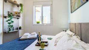 En snyggt bäddad säng med ett brickbord med champagne, glas och vindruvor, en handduksställning med prydligt vikta handdukar och ett fönster med utsikt över byggnader.
