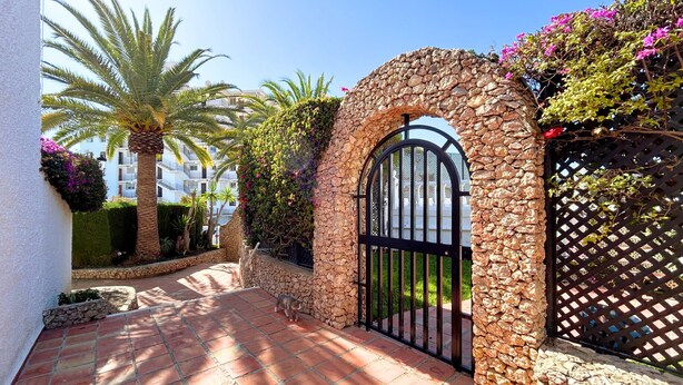 A stone gate with a metal arch, surrounded by lush greenery and a tiled pathway. - Verano Azul 72 (Nerja)