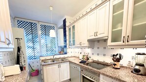 A kitchen with white cabinets, a granite countertop, a built-in oven, a sink, and a window with a lattice design. - Verano Azul 72 (Nerja)