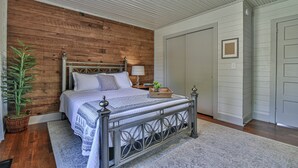 A bedroom with a wooden accent wall, a metal bed frame, a bedside table with a lamp, and a potted plant. - The Spruce House Cozy Home in Swannanoa (Swannanoa)