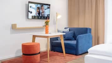 A modern room with a television mounted on the wall, a blue sofa, a small wooden table with a vase of flowers, and a red ottoman.