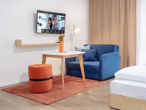 A modern room with a television mounted on the wall, a blue sofa, a small wooden table with a vase of flowers, and a red ottoman. - smartments Hamburg Hamm (Hamburg)