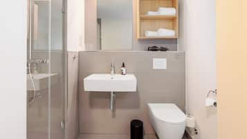 Un bagno moderno con box doccia in vetro, lavandino bianco, water e ripiano in legno con asciugamani.