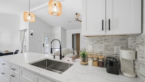 Een moderne keuken met witte kasten, een groot eiland met een spoelbak, hanglampen en een plafondventilator.