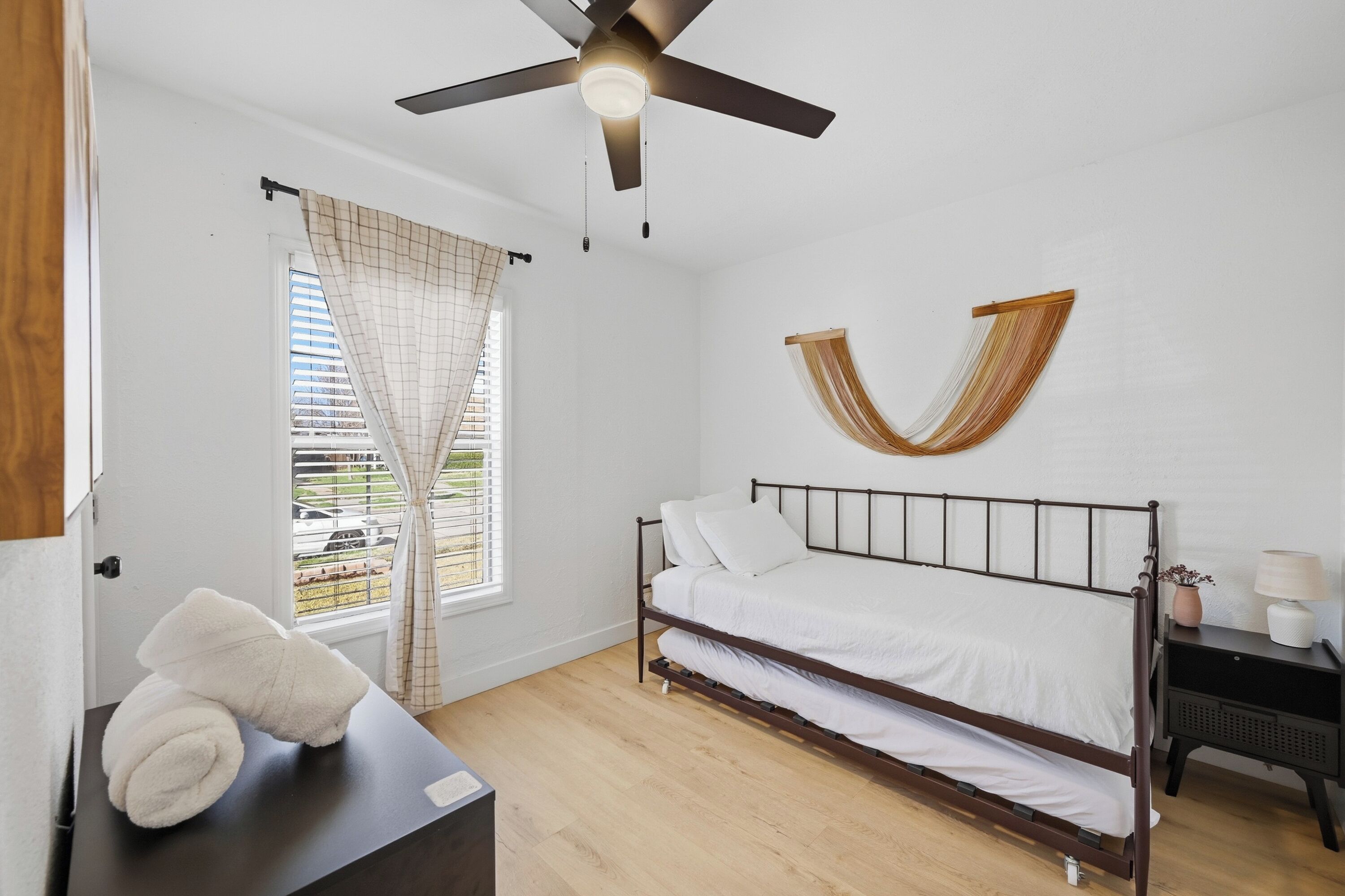 A bedroom with a bed, a ceiling fan, a window with curtains, a bedside table with a lamp, and a wall-mounted decorative item.
