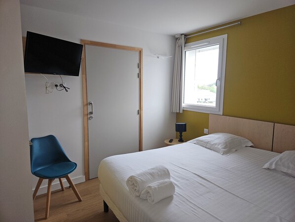 Quarto de hotel com cama, uma cadeira, televisão fixada na parede e janela com cortinas.