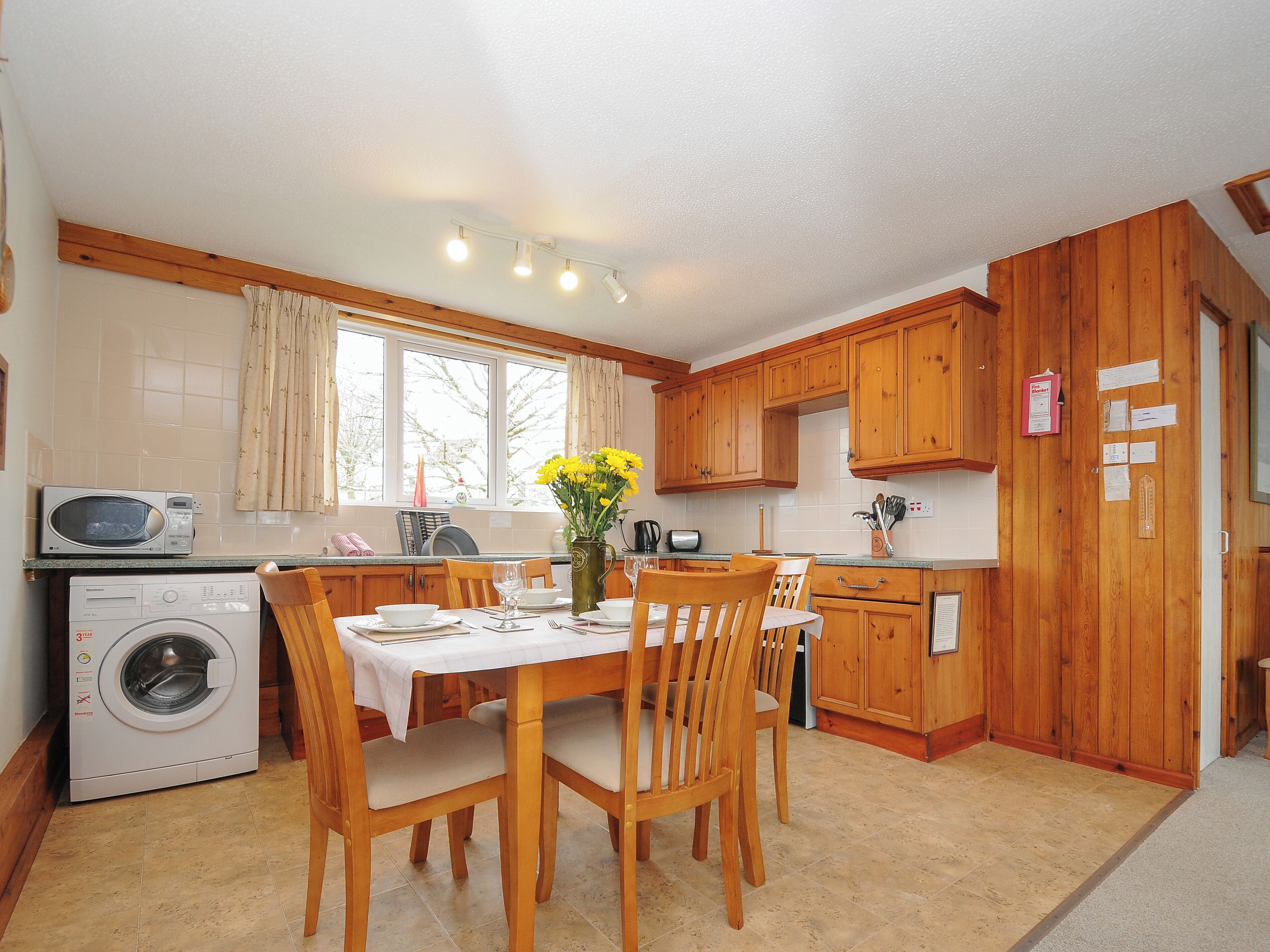 A compact kitchen with wooden cabinets, a washing machine, a microwave, and a dining area with a table set for two.